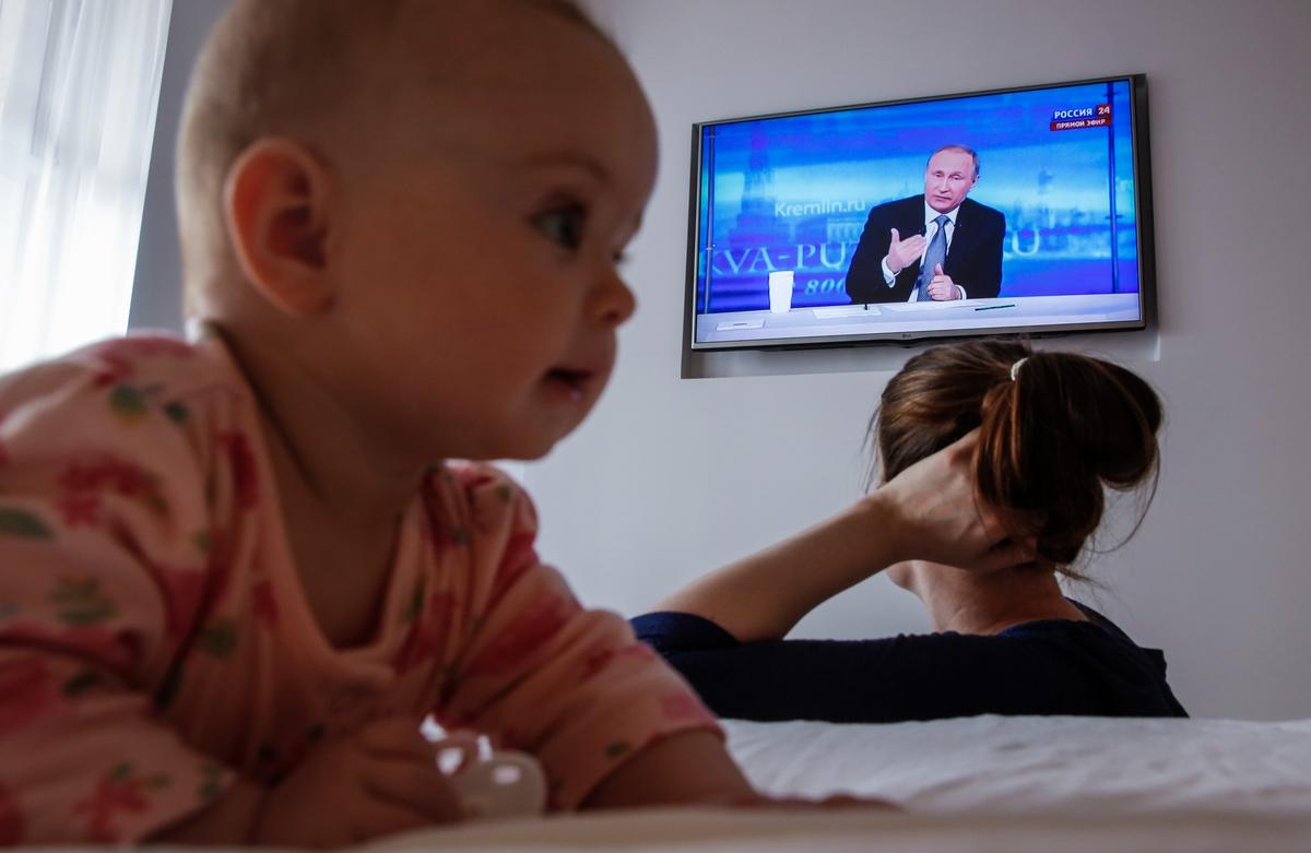 A baby crawls behind its mother as she watches Vladimir Putin take questions during his annual call-in show, in Moscow, 14 April 2016. Photo: EPA / SERGEI ILNITSKY